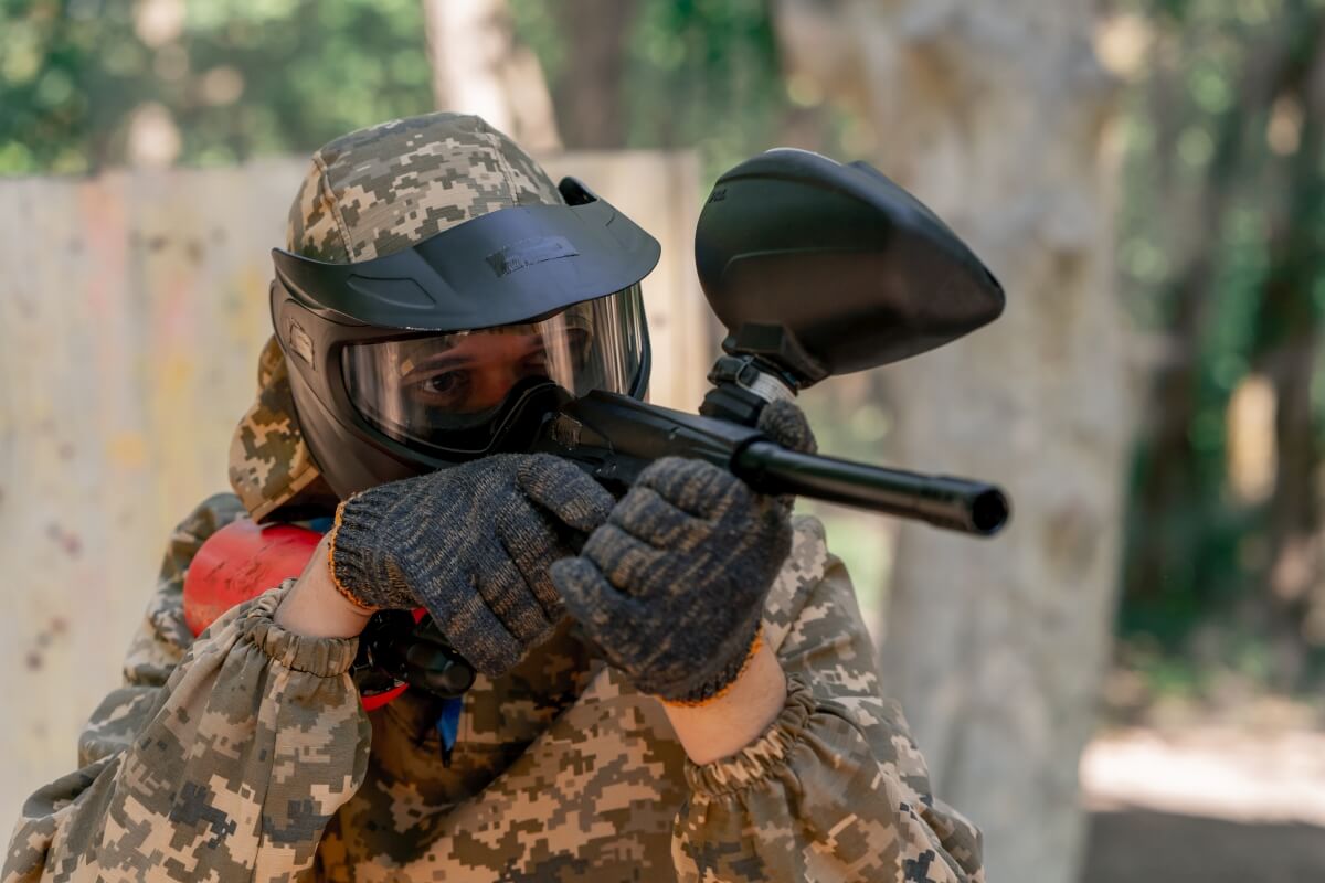 Osoba w maskującym ubraniu i zabezpieczającym hełmie celuje z markera paintballowego, paintball zabawa dla dzieci w odmianach.