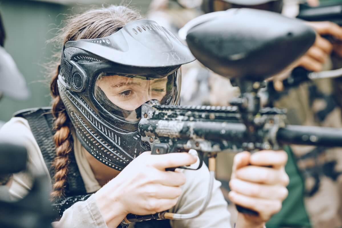 Kobieta w ochronnej masce i kamizelce celuje z markera na paintballowym wieczorze panieńskim.