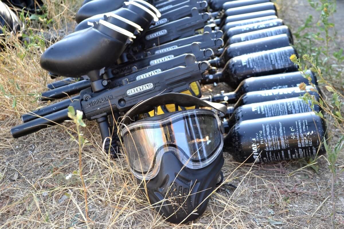 Wyposażenie do paintballu przygotowane dla dużej grupy uczestników, w tym markery i maski ochronne, zorganizowane na zewnątrz.