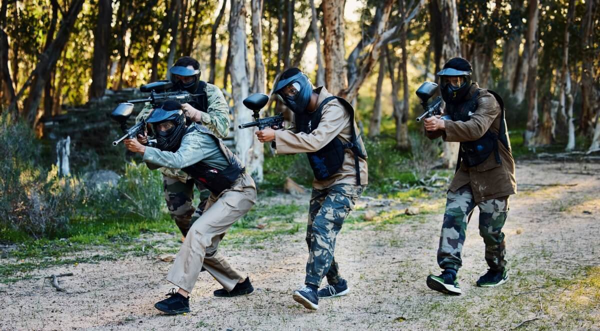 Grupa osób w maskach ochronnych bawi się w paintball, świętując urodziny dziecka.
