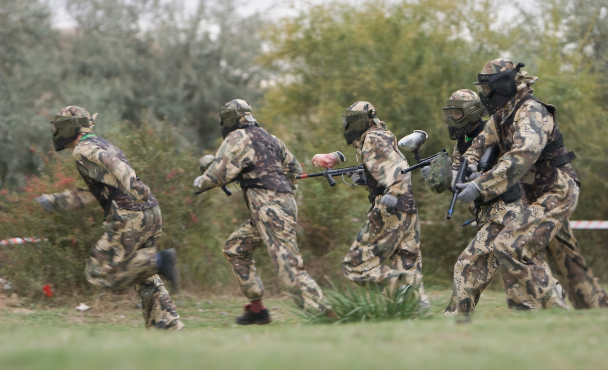 Grupa osób w mundurach biega po polu, uczestnicząc w scenariuszach paintball dla firm.