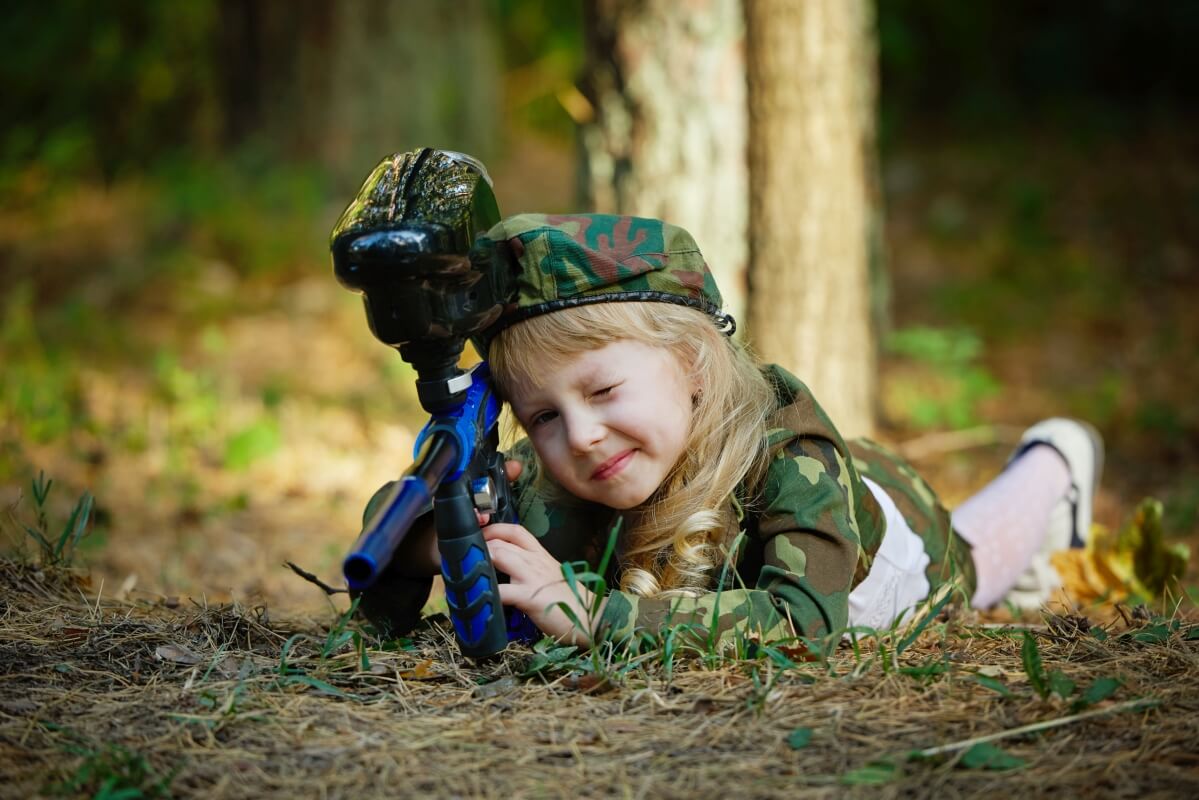 Dziewczynka w stroju kamuflażowym leży z markerem do paintballu, co zabrać paintballowe urodziny dziecka.