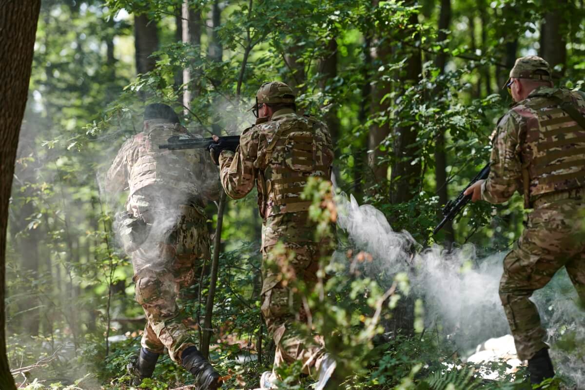 Trzech żołnierzy w wojskowych mundurach w lesie; przygotowanie dziecka do paintballa obejmuje naukę poruszania się w terenie.